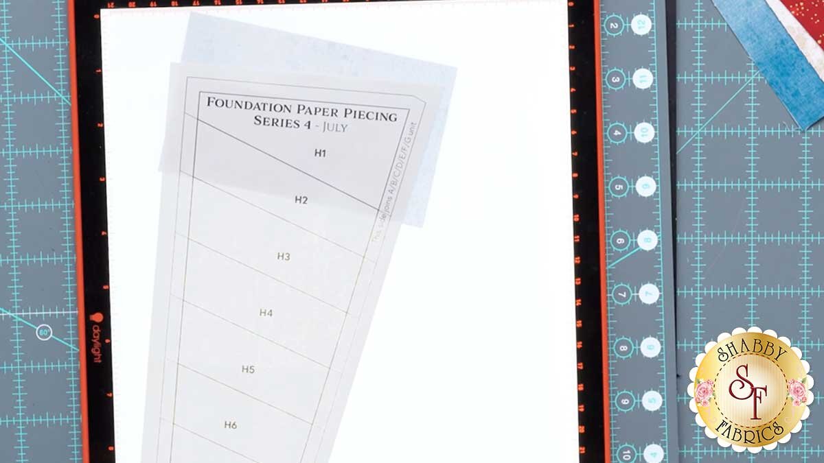 fpp_series_4_july_piecing_on_light_pad.jpg A foundation paper on a lit light pad with the first fabric piece positioned to cover its shape plus a quarter inch margin.