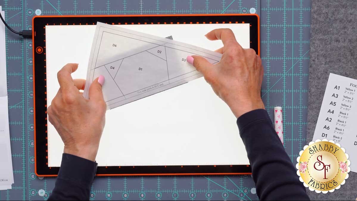 fpp_august_4_piecing_on_light_pad.jpg A foundation paper on a lit light pad showing the first fabric piece covering its shape with a quarter inch margin all around.