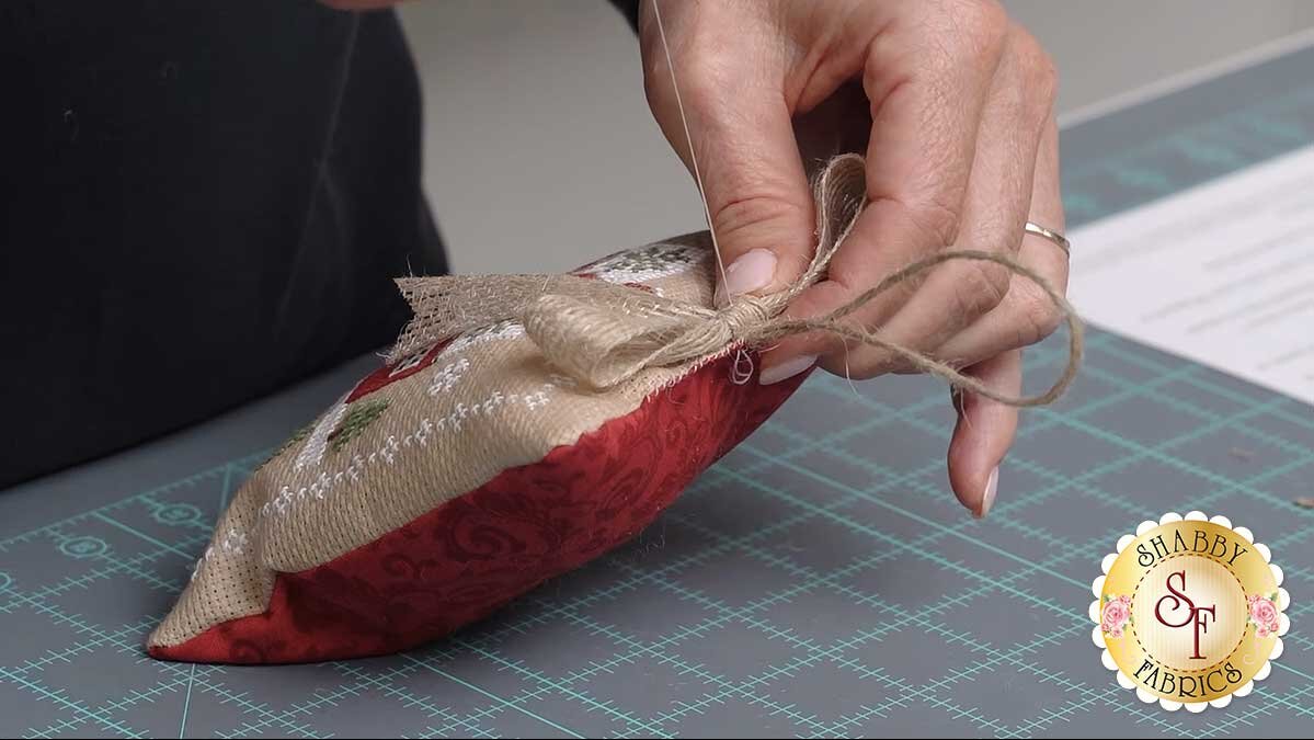 farmhouse_christmas_ornaments_stitching_the_bow.jpg Hands tying the ribbon into a bow, holding the center point and pushing the loop through.