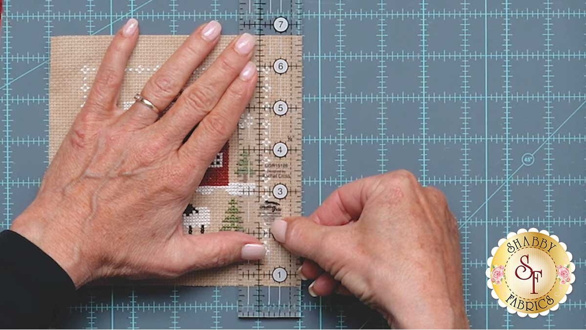 farmhouse_christmas_ornaments_measureing_quarter_inch.jpg A ruler positioned three quarters of an inch out from the outermost cross stitches, ready to trim the piece to size.